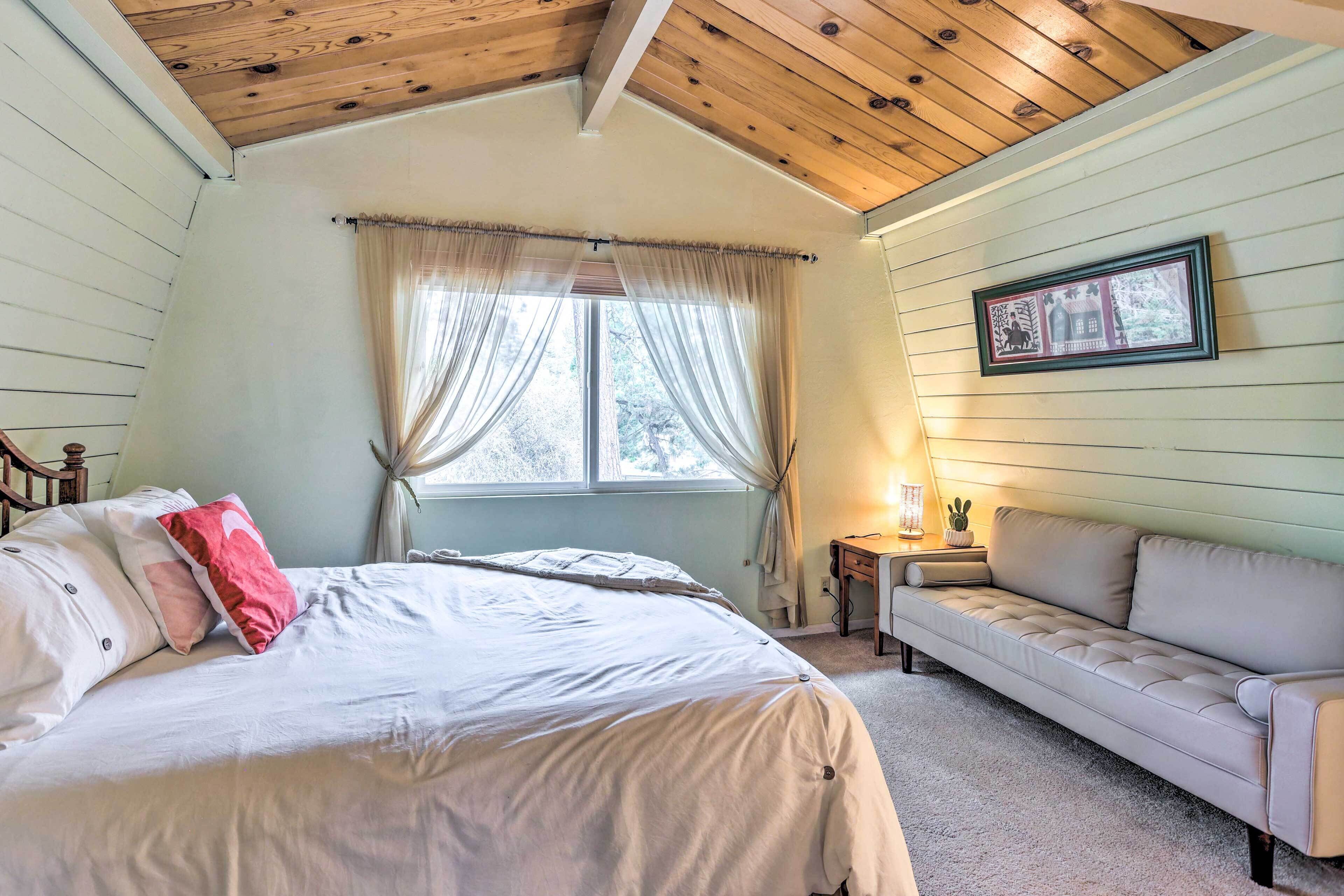 A bedroom with a bed, a sofa, a window with curtains, a framed picture on the wall, and a lamp on a side table.