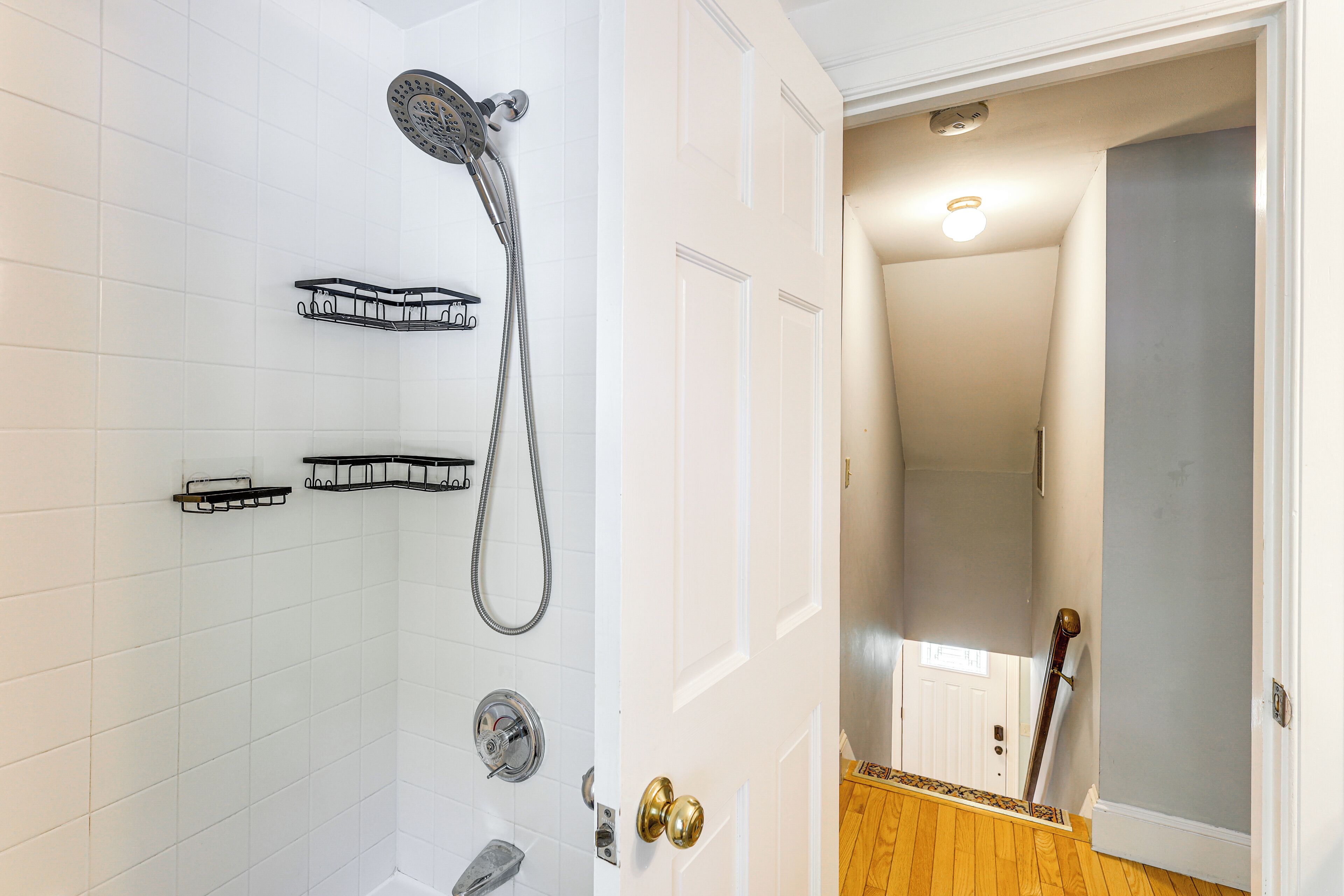 A bathroom with white tiled walls, a shower with a handheld showerhead, and a wooden floor.