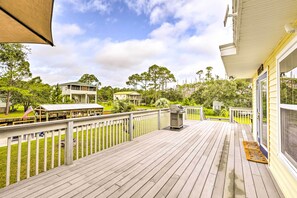 A wooden deck with a grill, a covered seating area, and a view of a green landscape with trees and a building in the background. - Waterfront Home: Spectacular On-site Fishing! (Panacea)