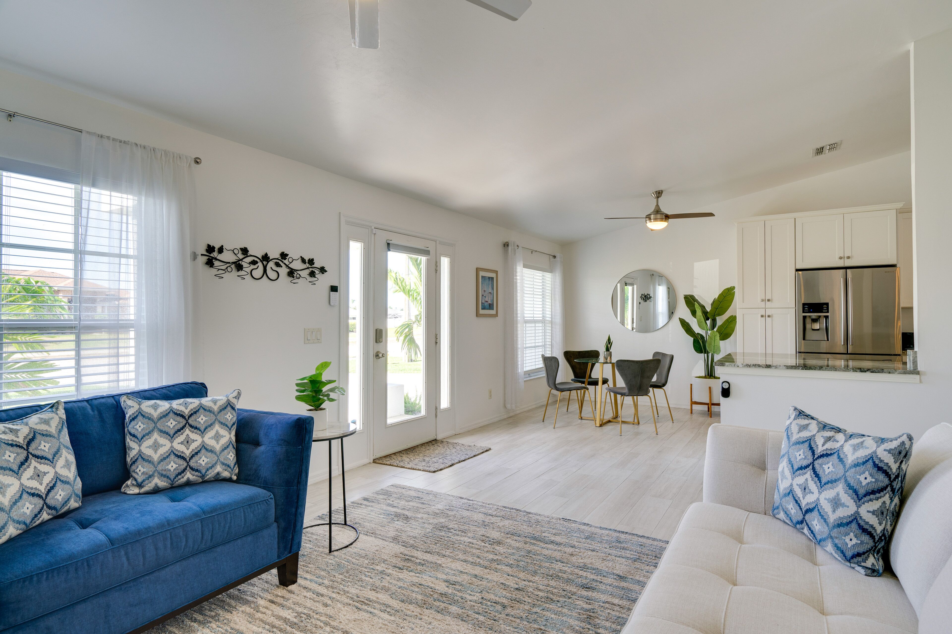 Un amplio salón con un sofá azul y un sofá blanco, una zona de comedor con mesa y sillas, y una cocina con armarios blancos y un refrigerador.