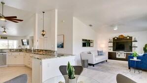 A modern kitchen with white cabinetry, a granite countertop, and a central island. There is a dining area with a glass table and chairs, and a living room with a blue sofa and a television. - Canal-front Cape Coral Home Rental: Pool, Lanai! (Cape Coral)
