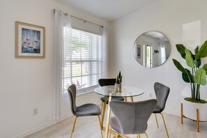 A dining area with a glass table, four grey chairs, a potted plant, and a framed painting on the wall.