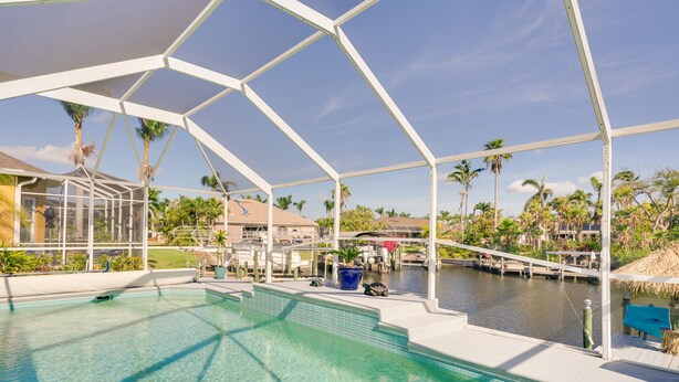 A pool area with a clear blue pool, a white canopy structure, and a view of a marina with boats and palm trees. - Canal-front Cape Coral Home Rental: Pool, Lanai! (Cape Coral)