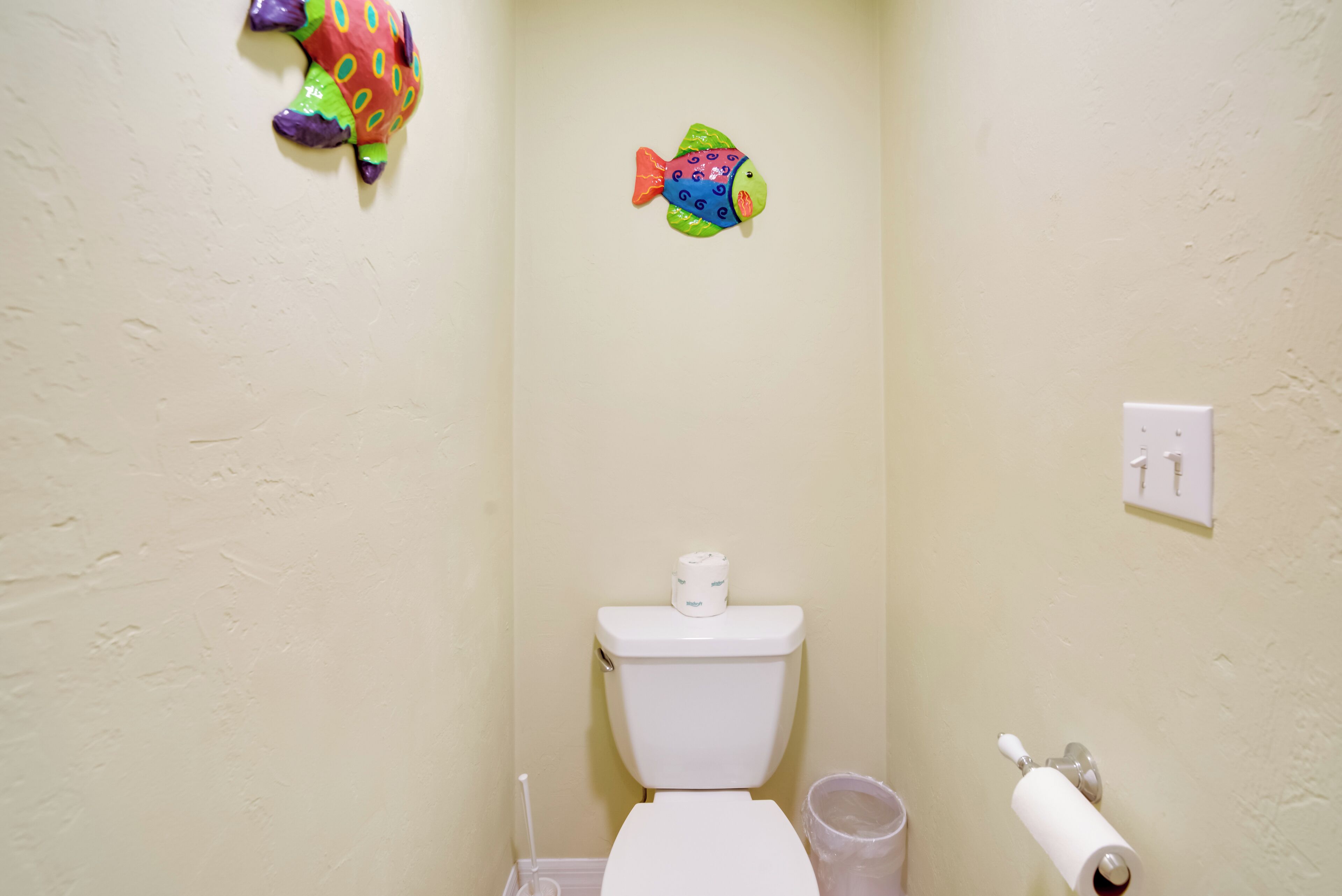 A bathroom with a toilet, a trash bin, a toilet paper holder, and a wall decoration.