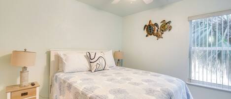 A bedroom with a bed, two bedside lamps, a ceiling fan, a window with a view, and a wall decoration.
