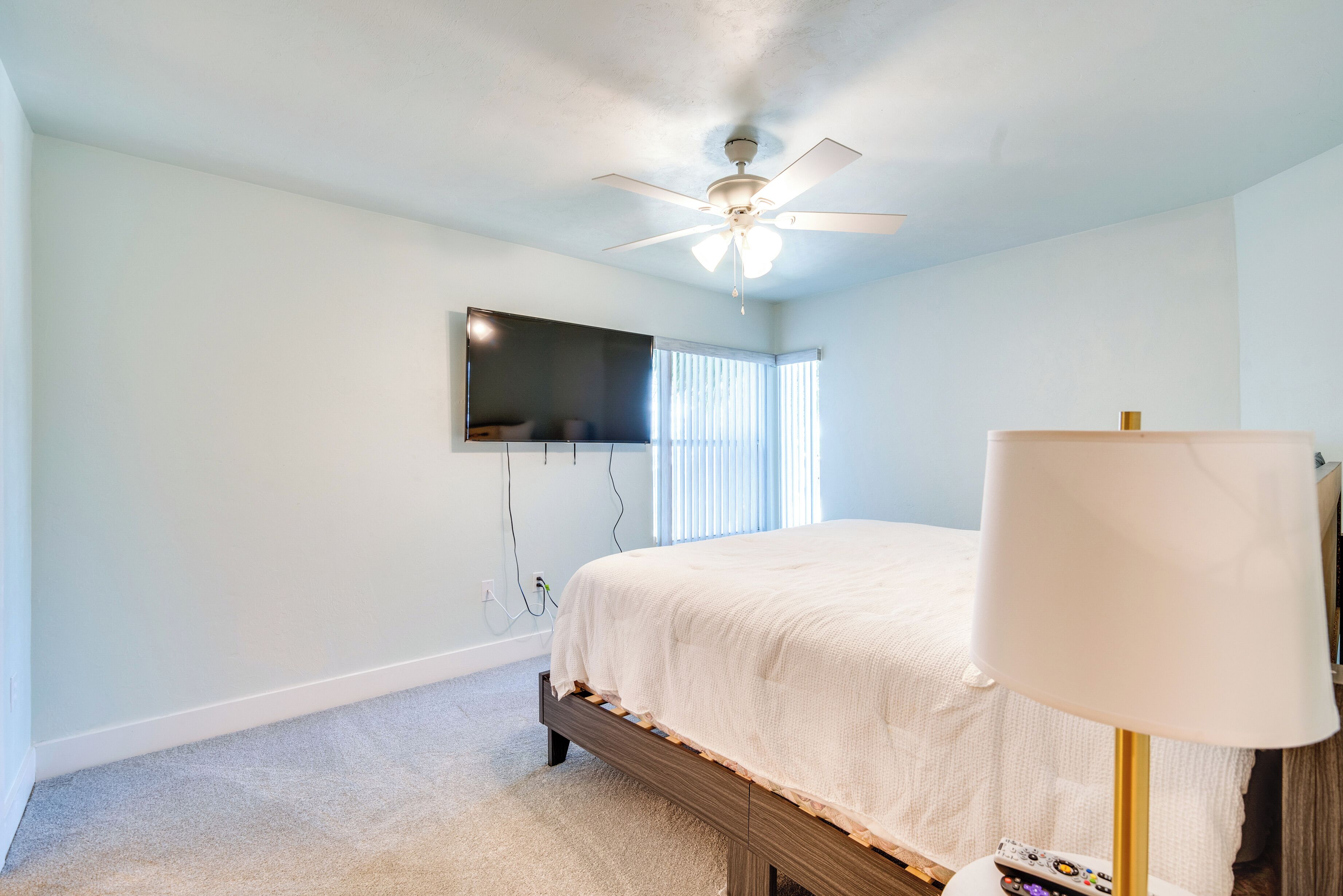 A bedroom with a bed, a ceiling fan, a TV mounted on the wall, and a lamp.