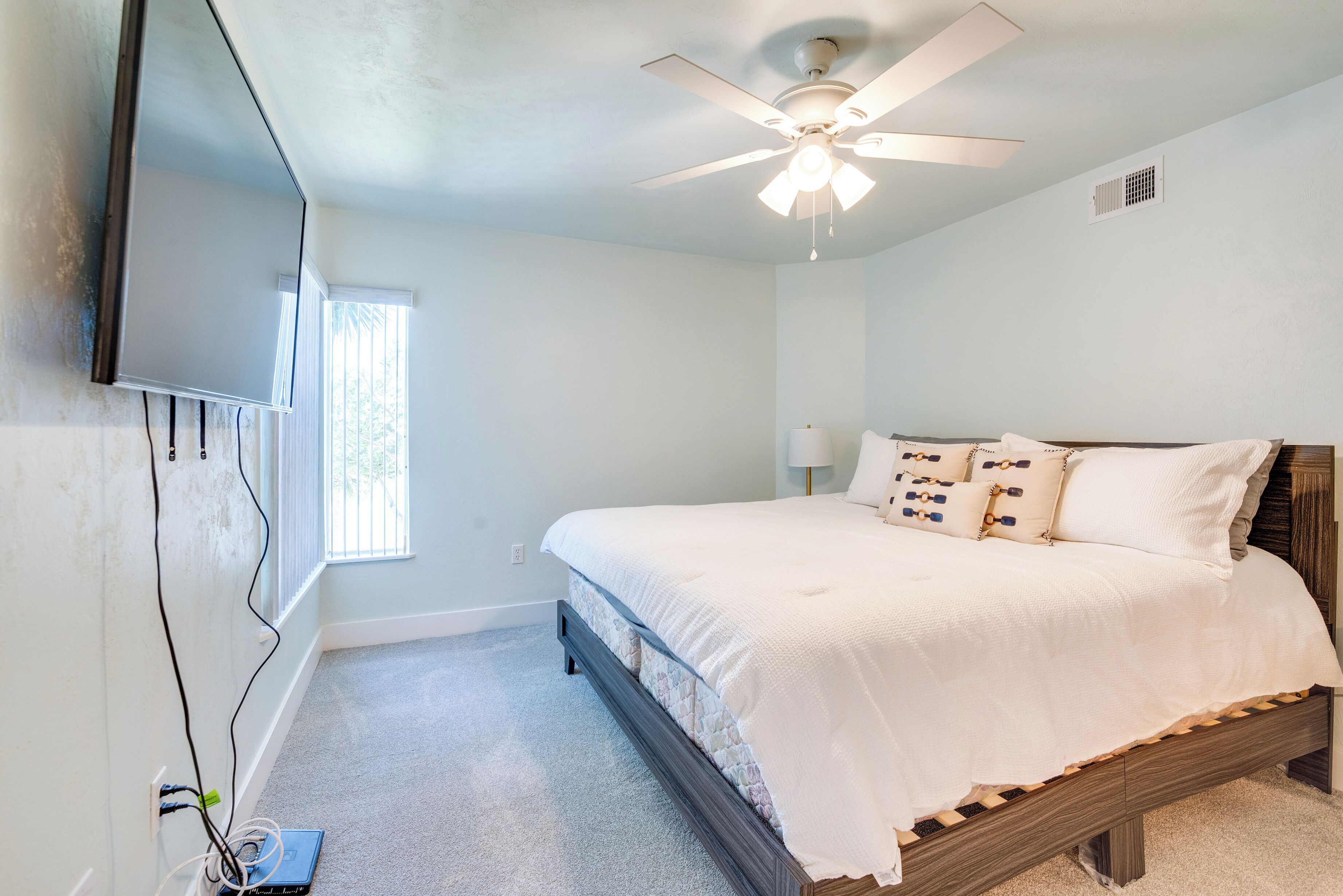 A bedroom with a bed, a ceiling fan, a TV mounted on the wall, and a window.
