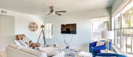 A living room with a beige sofa, a blue armchair, a ceiling fan, a mounted TV, and a large window with a view of palm trees.
