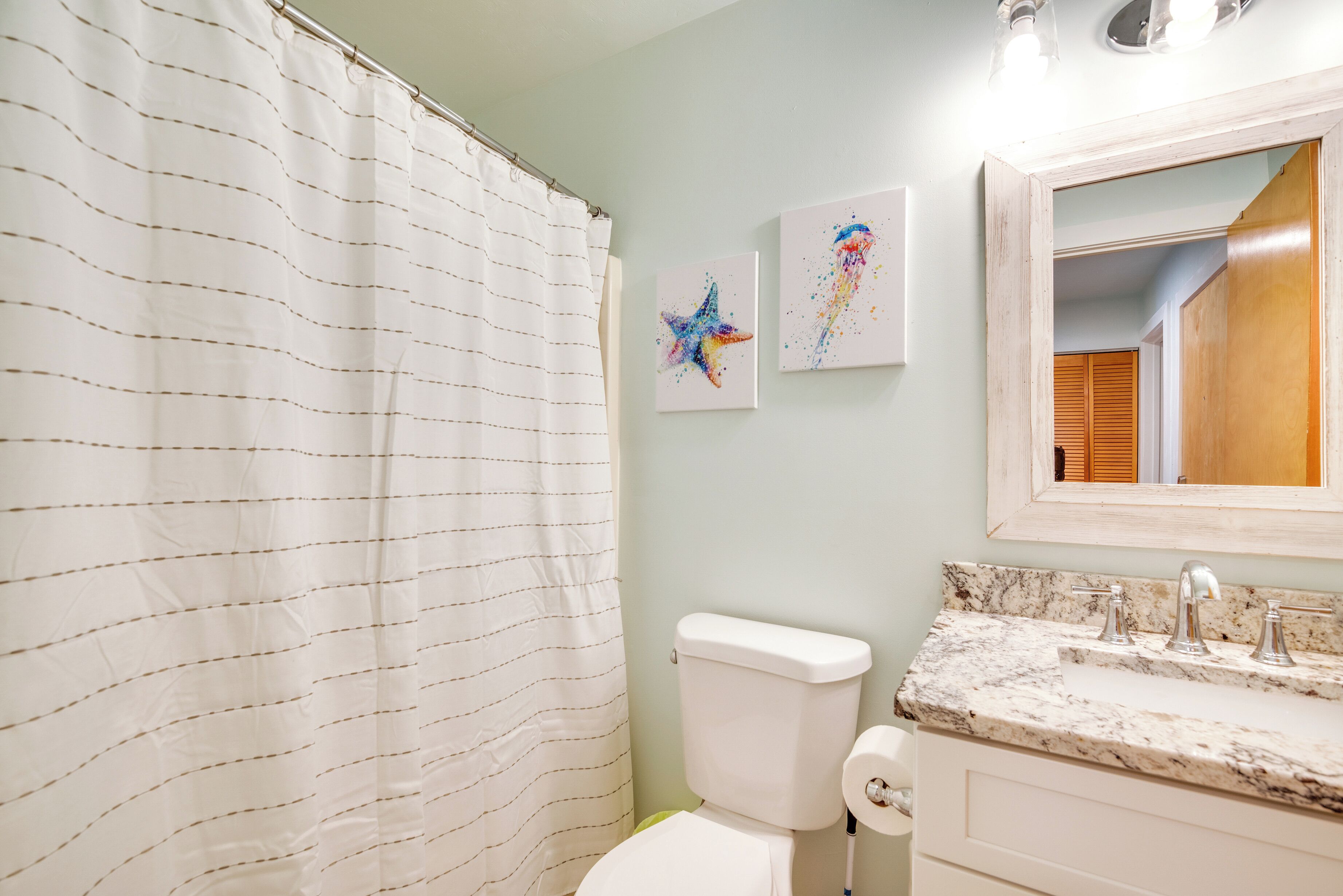 A bathroom with a striped shower curtain, a toilet, a sink with a marble countertop, and two wall-mounted light fixtures.