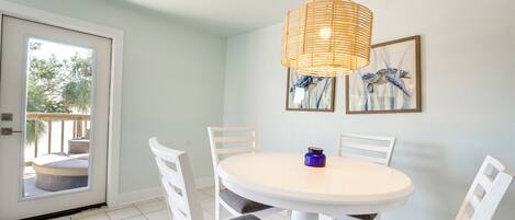 Un espace repas avec une table ronde, des chaises blanches, un luminaire suspendu en osier et une porte donnant sur un coin salon extérieur.
