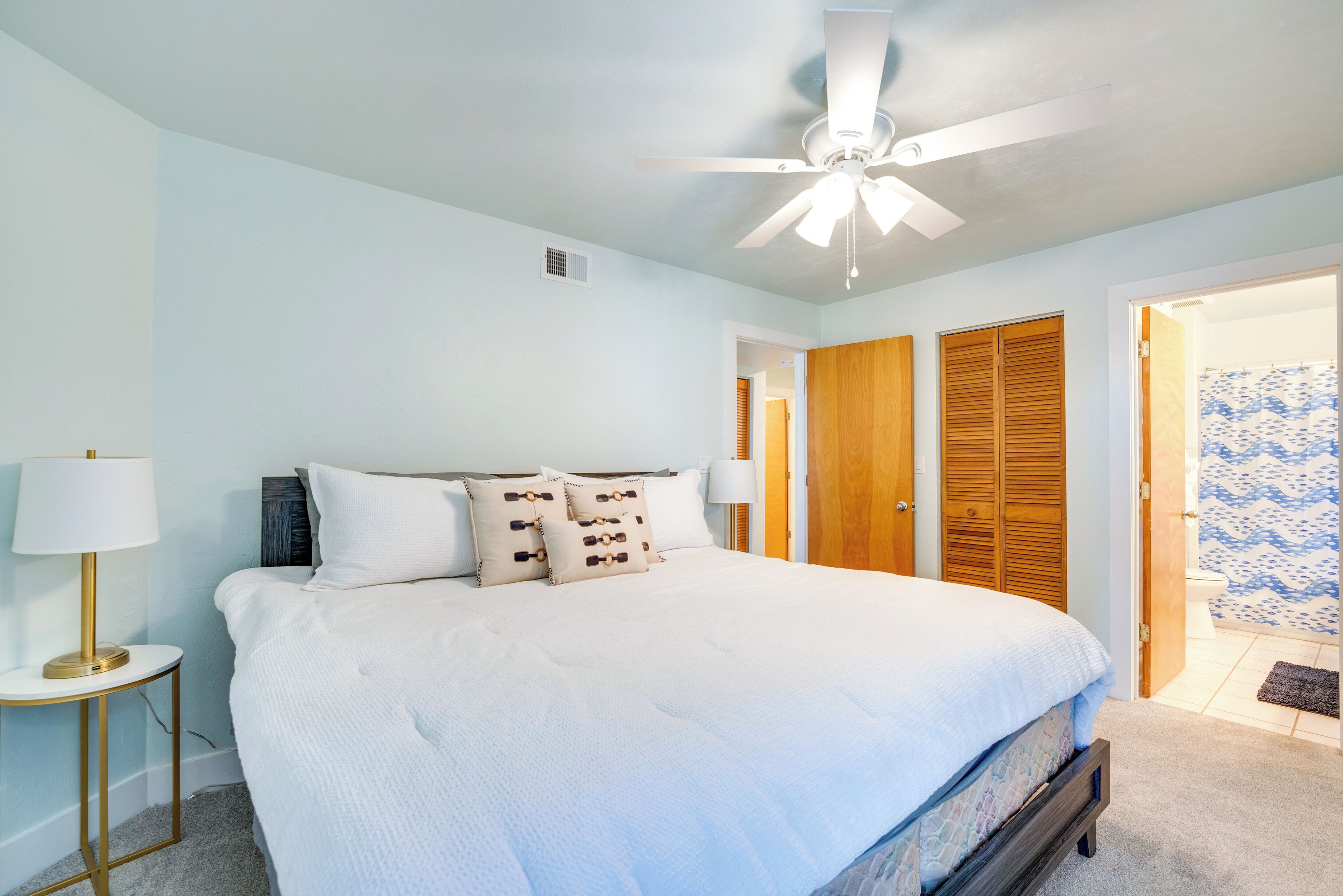 Quarto com cama, luminárias de cabeceira, ventilador de teto e portas de madeira.