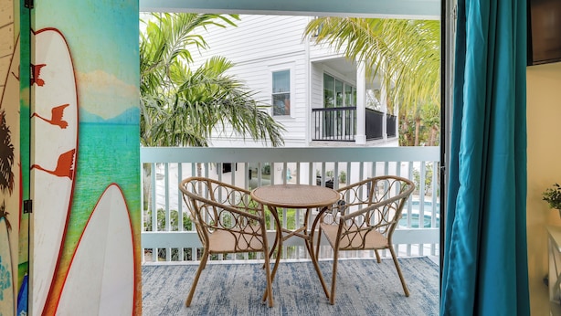 Een balkon met rieten stoelen en een tafeltje, met uitzicht op een wit huis met een zwembad en palmbomen.