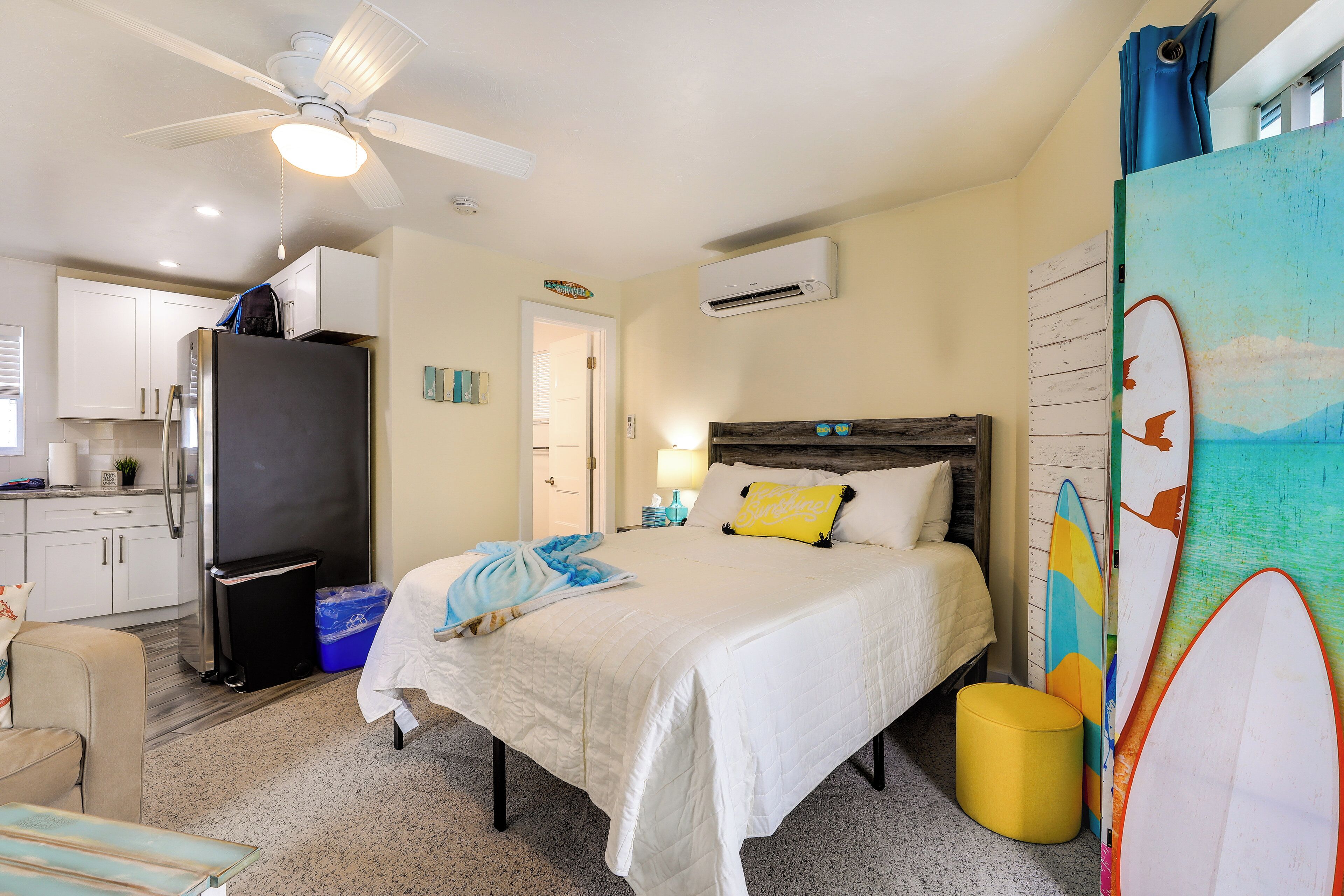 Quarto com cama, ventilador de teto, ar-condicionado, um banquinho pequeno e pranchas de surf encostadas na parede.