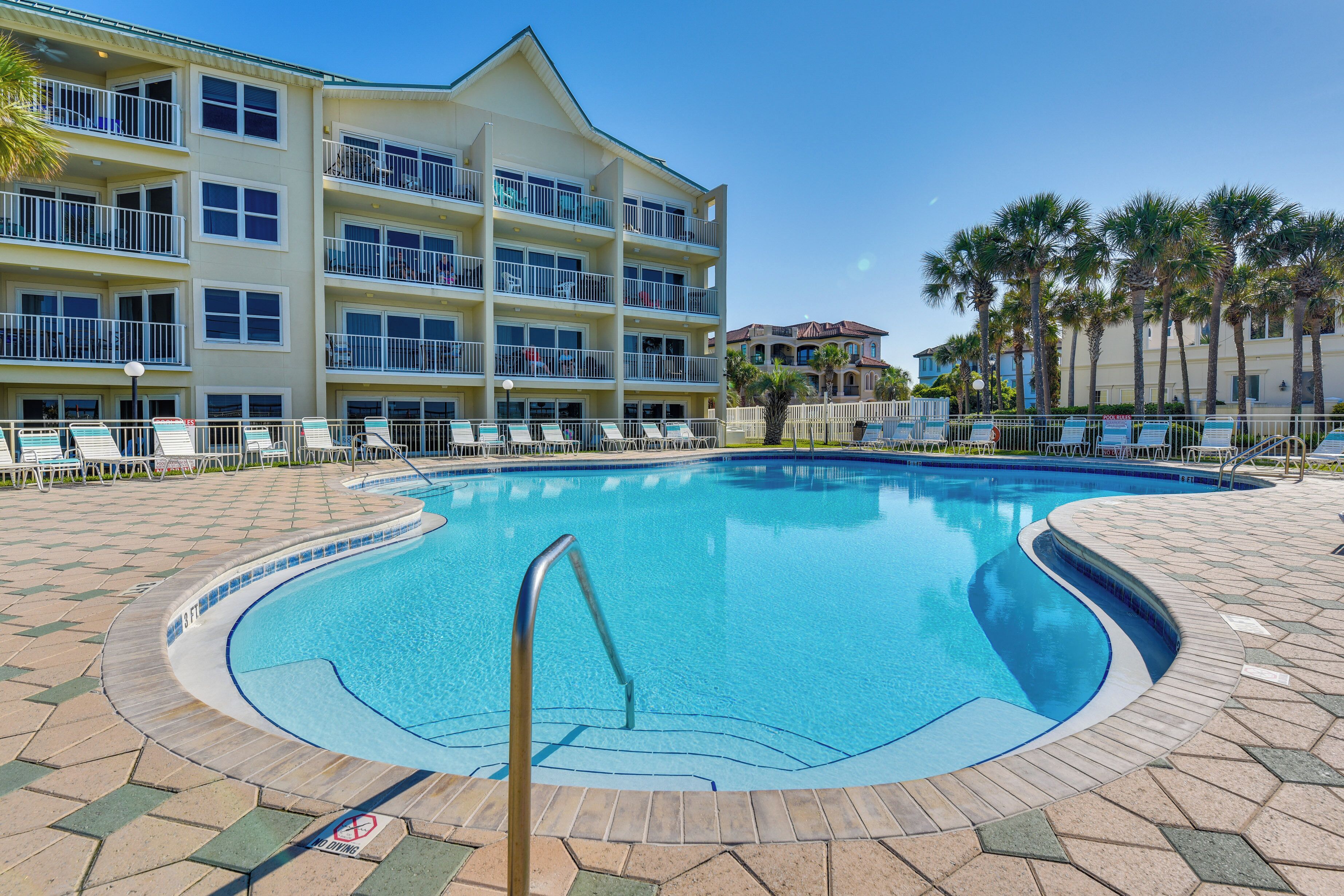 A swimming pool with a tiled deck, surrounded by lounge chairs and a multi-story building with balconies.
