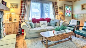 Un intérieur de chalet chaleureux avec un plafond en bois, un canapé agrémenté d’un coussin sur lequel est brodé le mot « Rassemblement », une table basse en bois et une fenêtre décorée d’un rideau à carreaux.