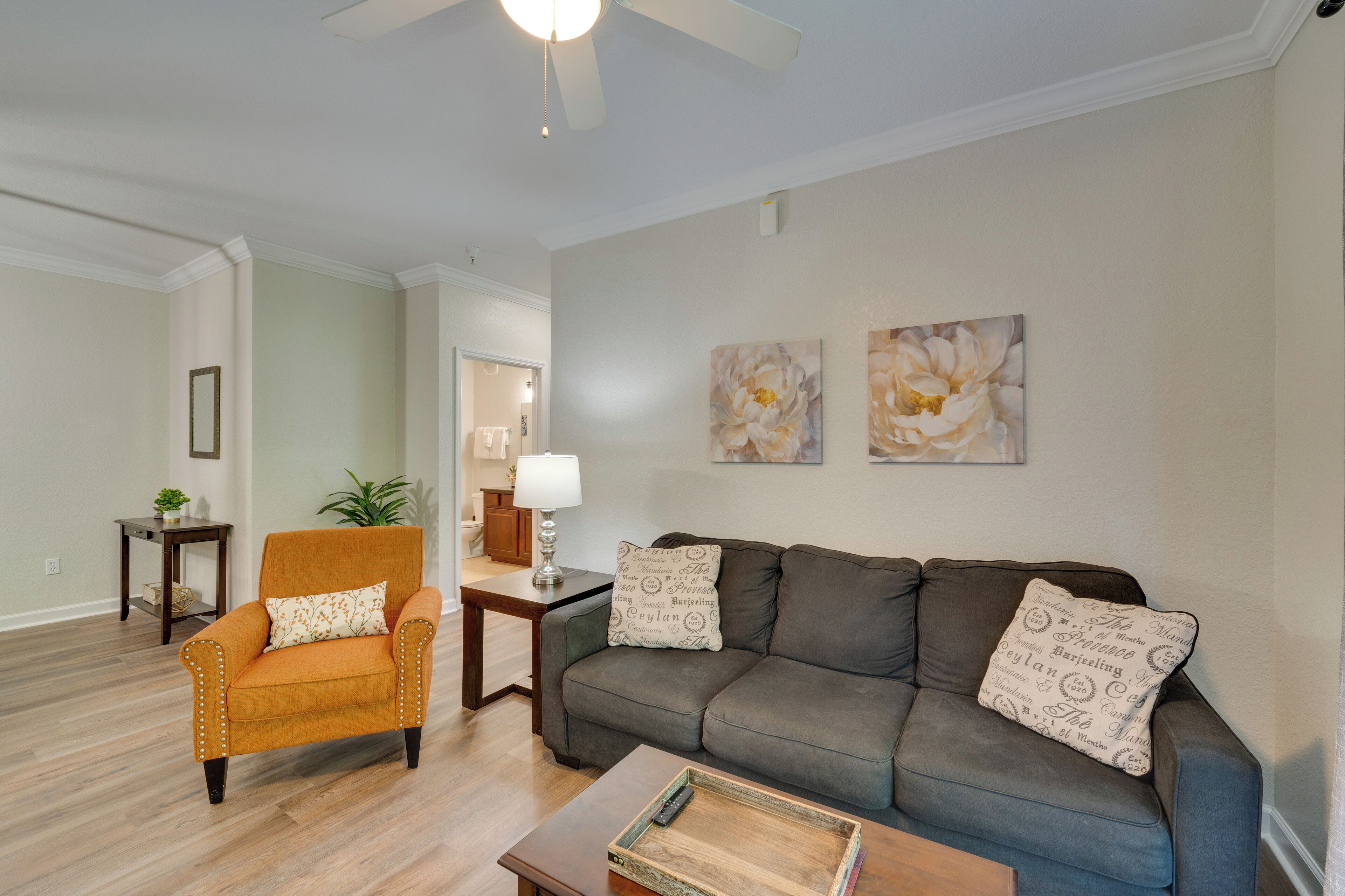 A living room with a sofa, armchair, and coffee table. There are two wall-mounted paintings and a ceiling fan.