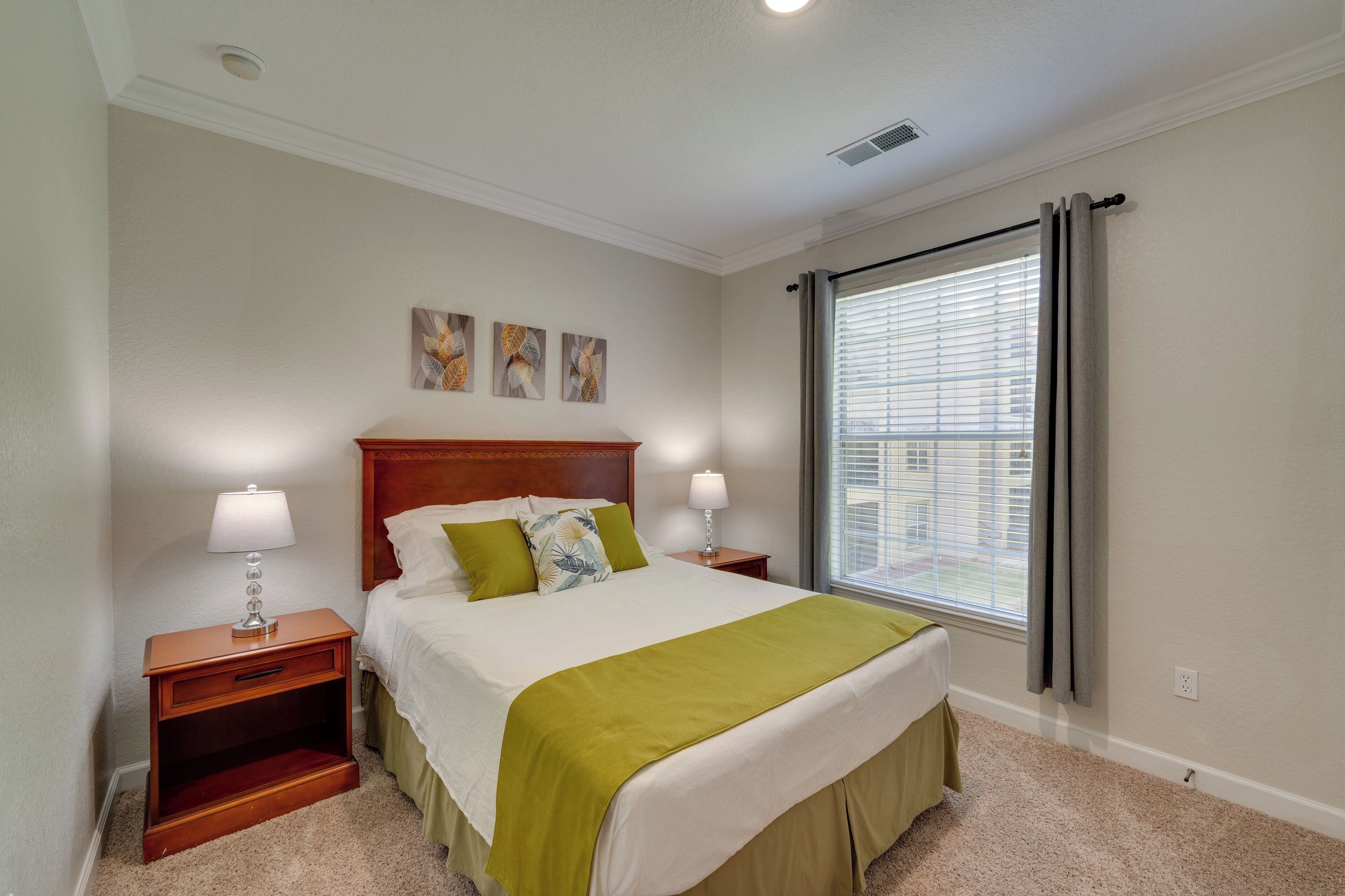 Une chambre à coucher bien aménagée, avec un grand lit, deux tables de chevet, une commode, une fenêtre avec des stores et trois œuvres d’art accrochées au mur.