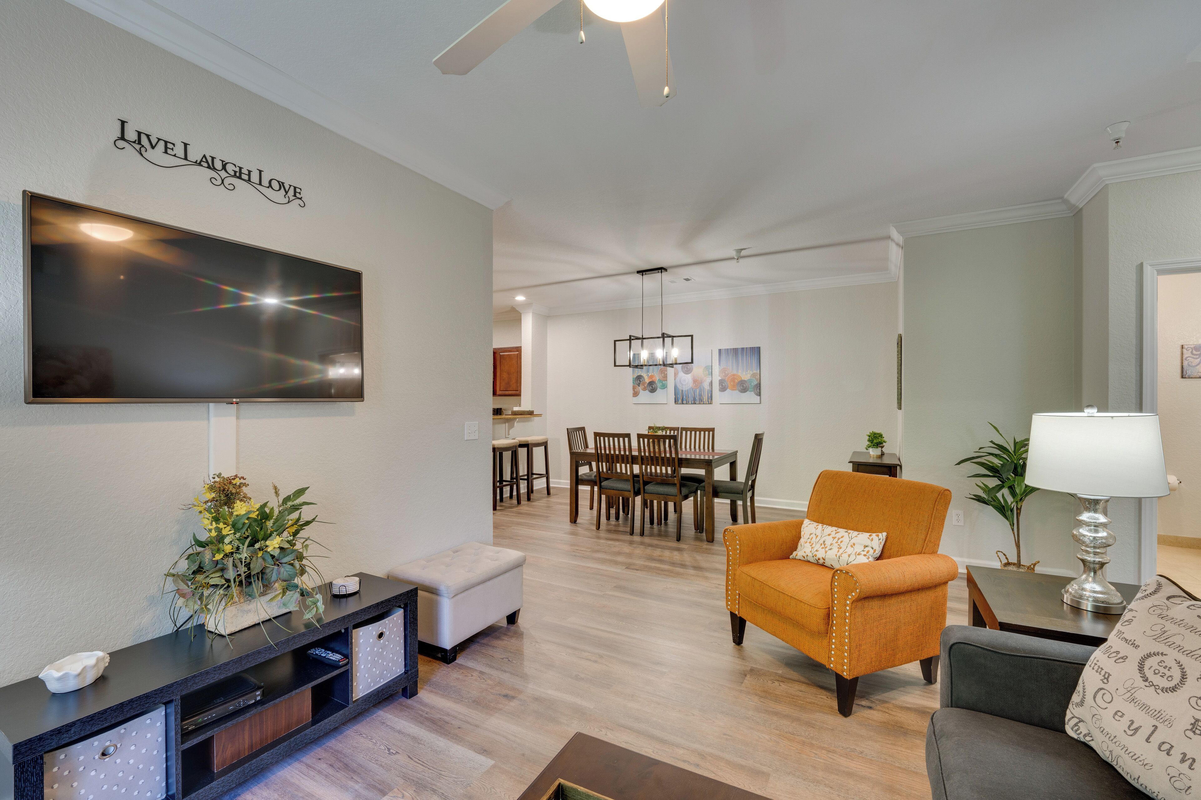 A modern living room with a flat-screen TV, a dining area with wooden furniture, and a cozy seating area with an orange armchair and a grey sofa.