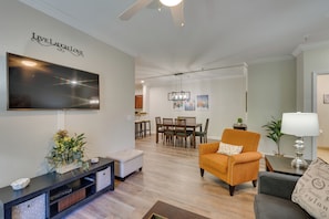 A modern living room with a flat-screen TV, a dining area with wooden chairs, and a cozy orange armchair.