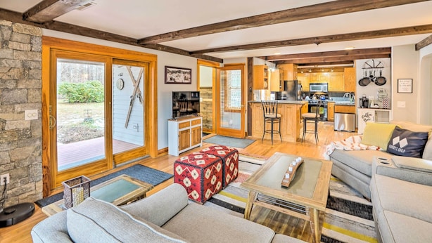 Una sala de estar acogedora con sofá, reposapiés y mesa de centro, una cocina con alacenas de madera y un comedor, y ventanas amplias con vistas a zonas verdes.