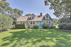 A two-story house with a blue front door, surrounded by a well-maintained lawn and various shrubs. - Cape Cod Cottage w/ Deck, Grill, & Games! (Falmouth)