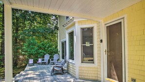 A covered porch with a wooden deck, white chairs, and a door with a screen.