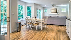 A dining area with a wooden table and chairs, a ceiling fan, and large windows offering a view of greenery.