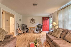 A living room with a wooden floor, two sofas, a guitar, a wall clock, and a red door. - West Yarmouth Home w/ Deck < 2 Mi to Beach! (West Yarmouth)