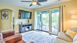 A spacious living room with a ceiling fan, a large window with curtains, a sofa, a coffee table, and a TV mounted on the wall.