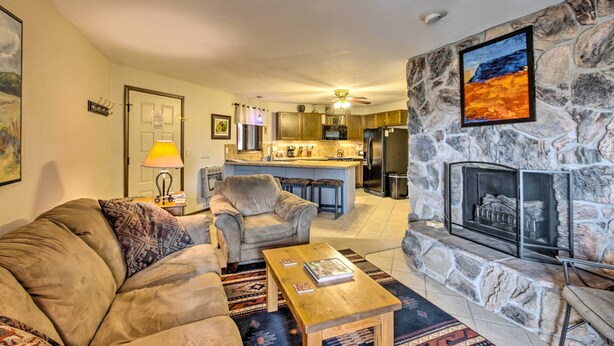 A living room with a sofa, coffee table, and fireplace. There is a kitchen in the background with a stone wall and a painting. - New Mexico Condo w/ Fireplace & Mountain View (Angel Fire)