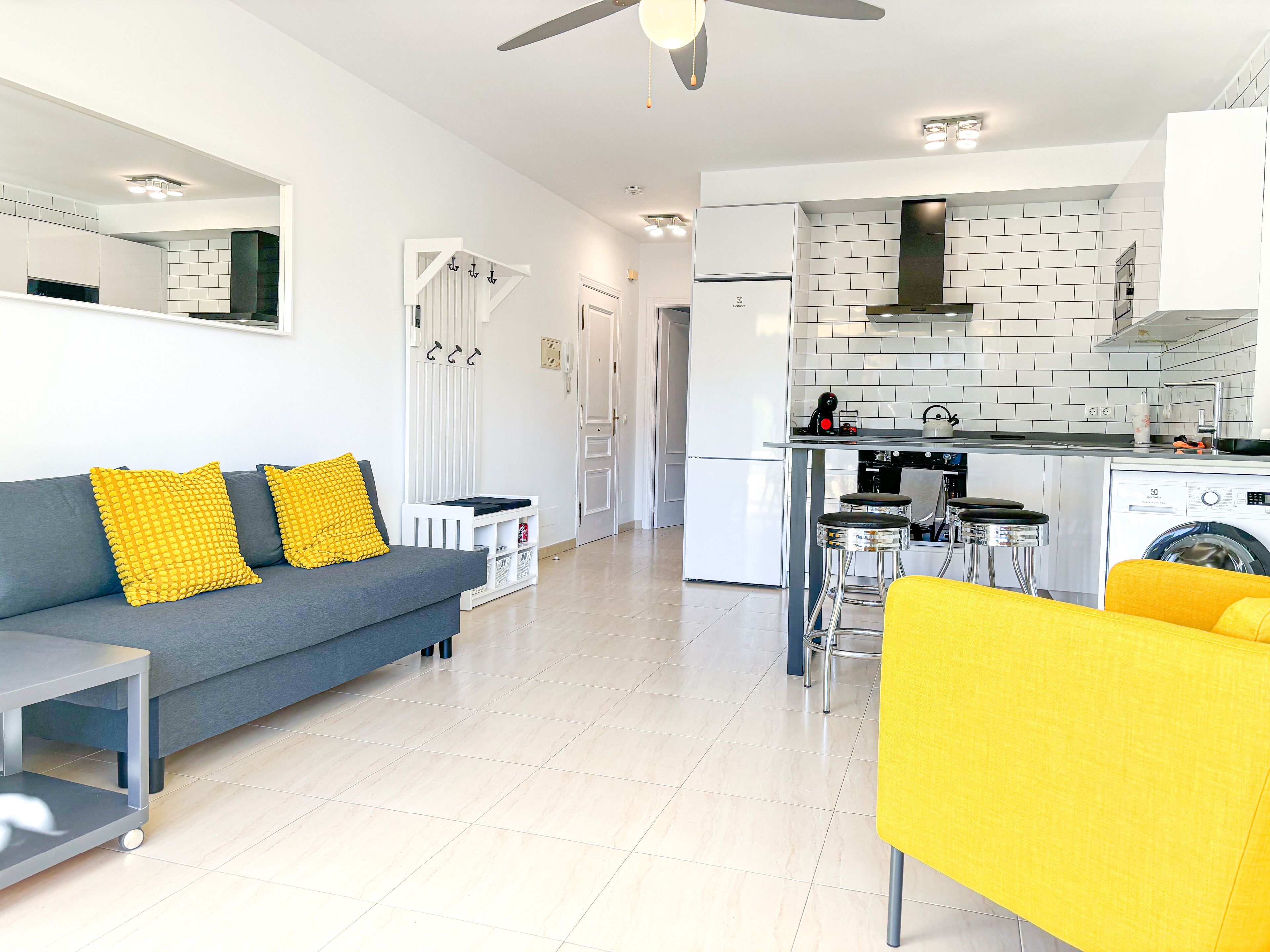 Un appartement moderne avec un canapé gris, des coussins jaunes, une petite table blanche, une cuisine équipée d’un lave-linge et un ventilateur de plafond.