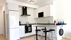 Een moderne keuken met witte kasten, een ingebouwde oven, een zwarte afzuigkap en een roestvrijstalen spoelbak. Drie barkrukken en een wasmachine zijn aanwezig.