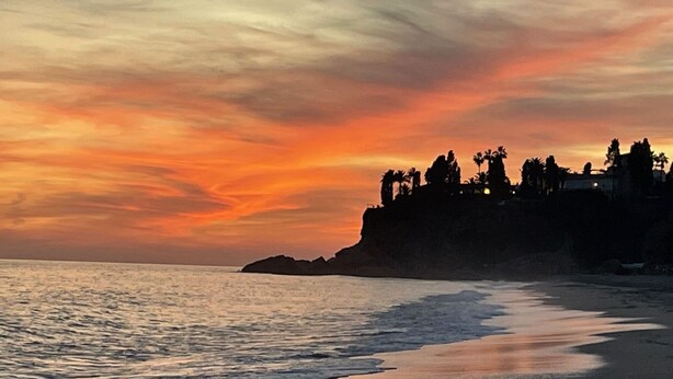Een strand met een zonsondergang, een rustige zee en een rotsachtige klif met bomen.