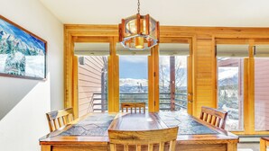 A wooden dining table with six chairs, a hanging light fixture, and large windows with a mountain view.