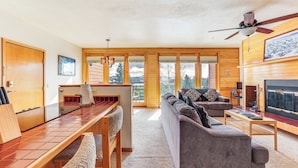 A modern living room with a sofa, coffee table, and a fireplace. There is a dining area with a table and chairs, and a kitchen visible through an open door.