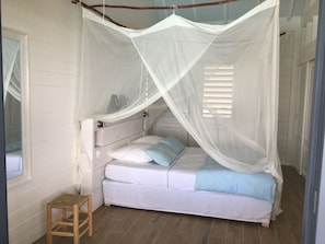 A single bed with a canopy, a small wooden stool, and a window with blinds.