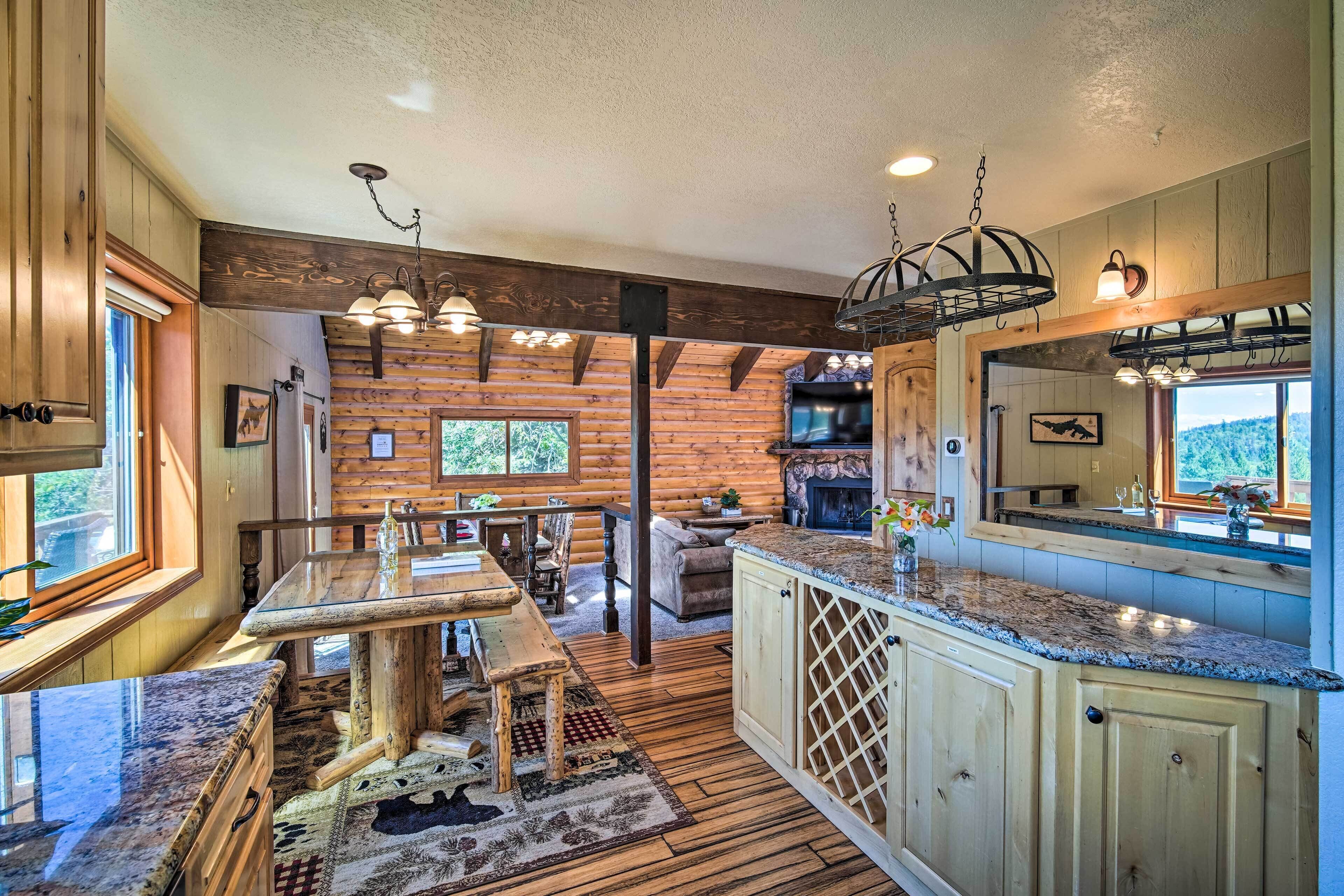 A rustic cabin interior with a wooden dining area, a kitchen with granite countertops, and a large window overlooking a scenic landscape.