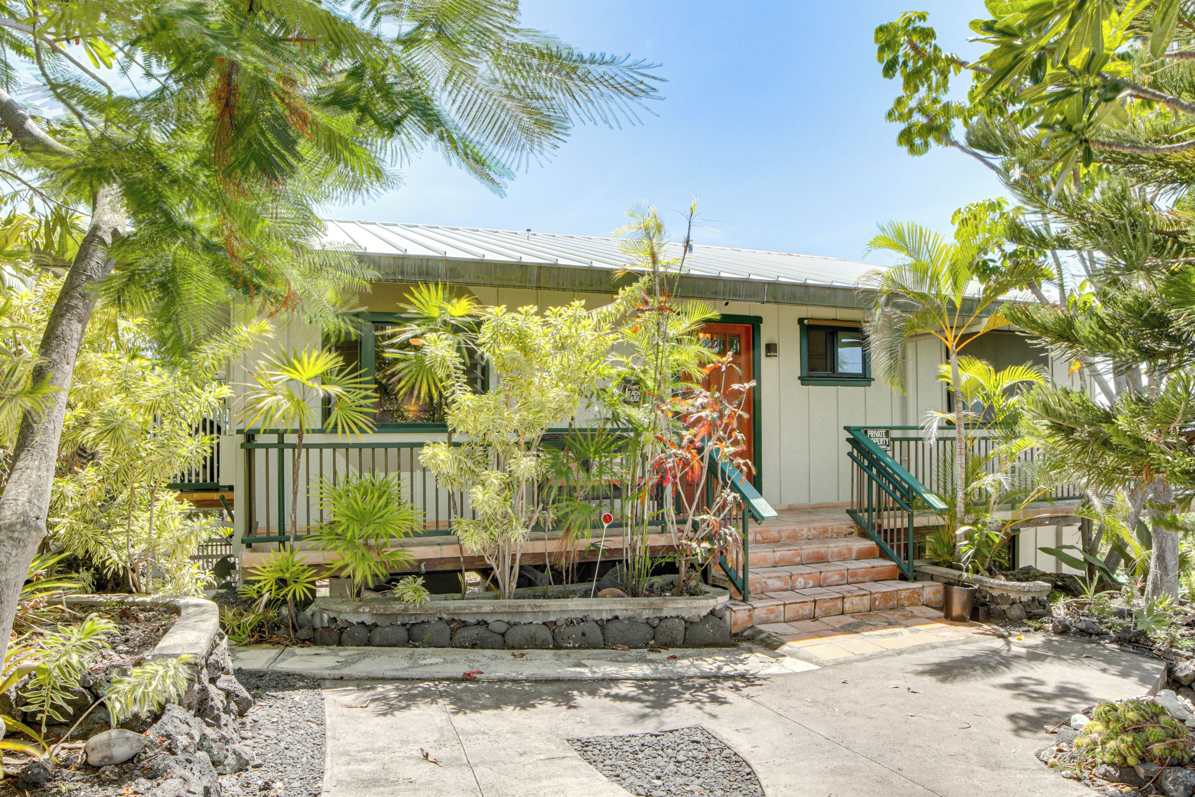 A house with a stone pathway leading to a wooden deck, surrounded by tropical plants and a metal staircase.