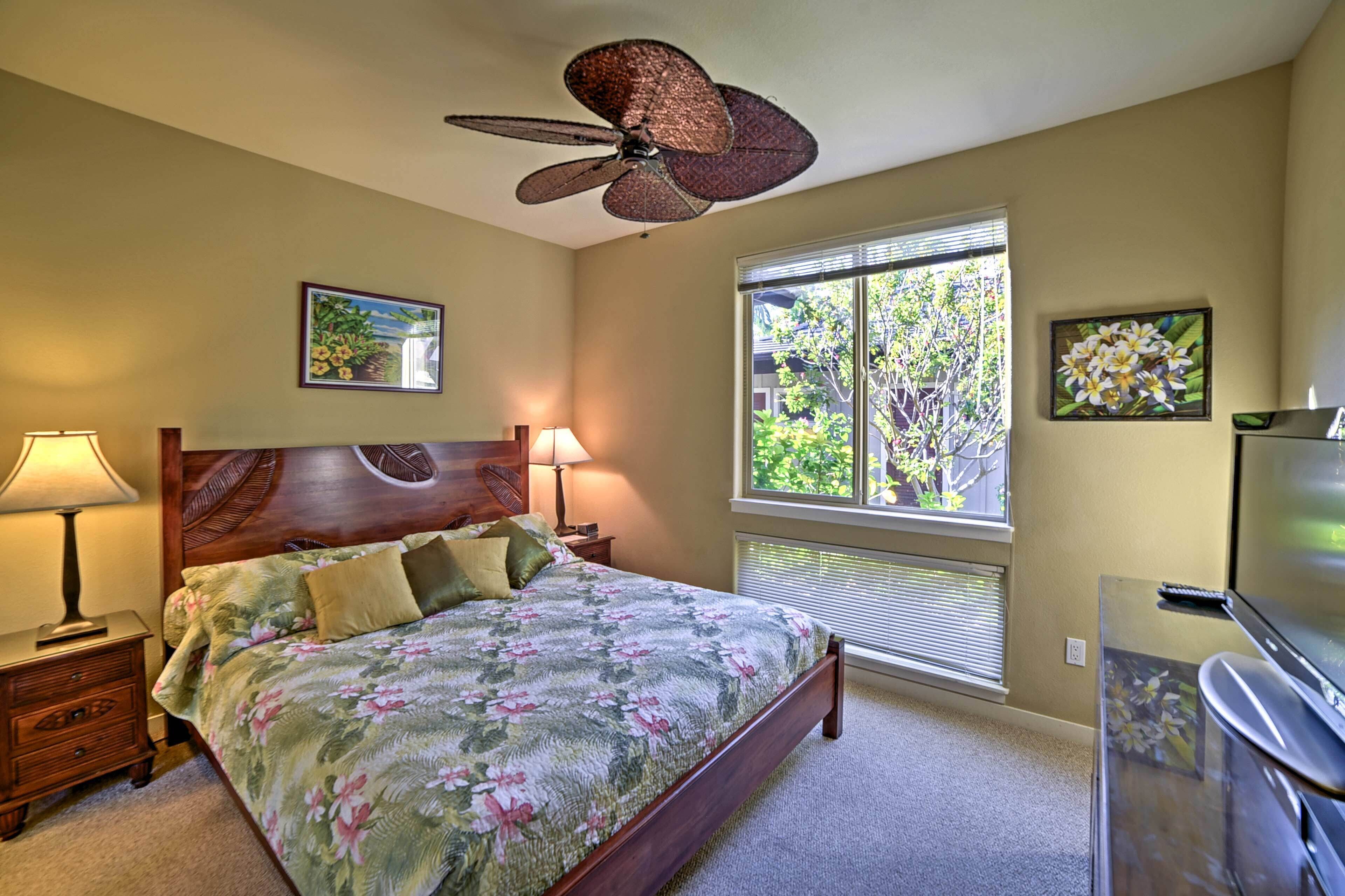 A bedroom with a wooden bed, two bedside lamps, a ceiling fan, a large window with blinds, and a TV stand.