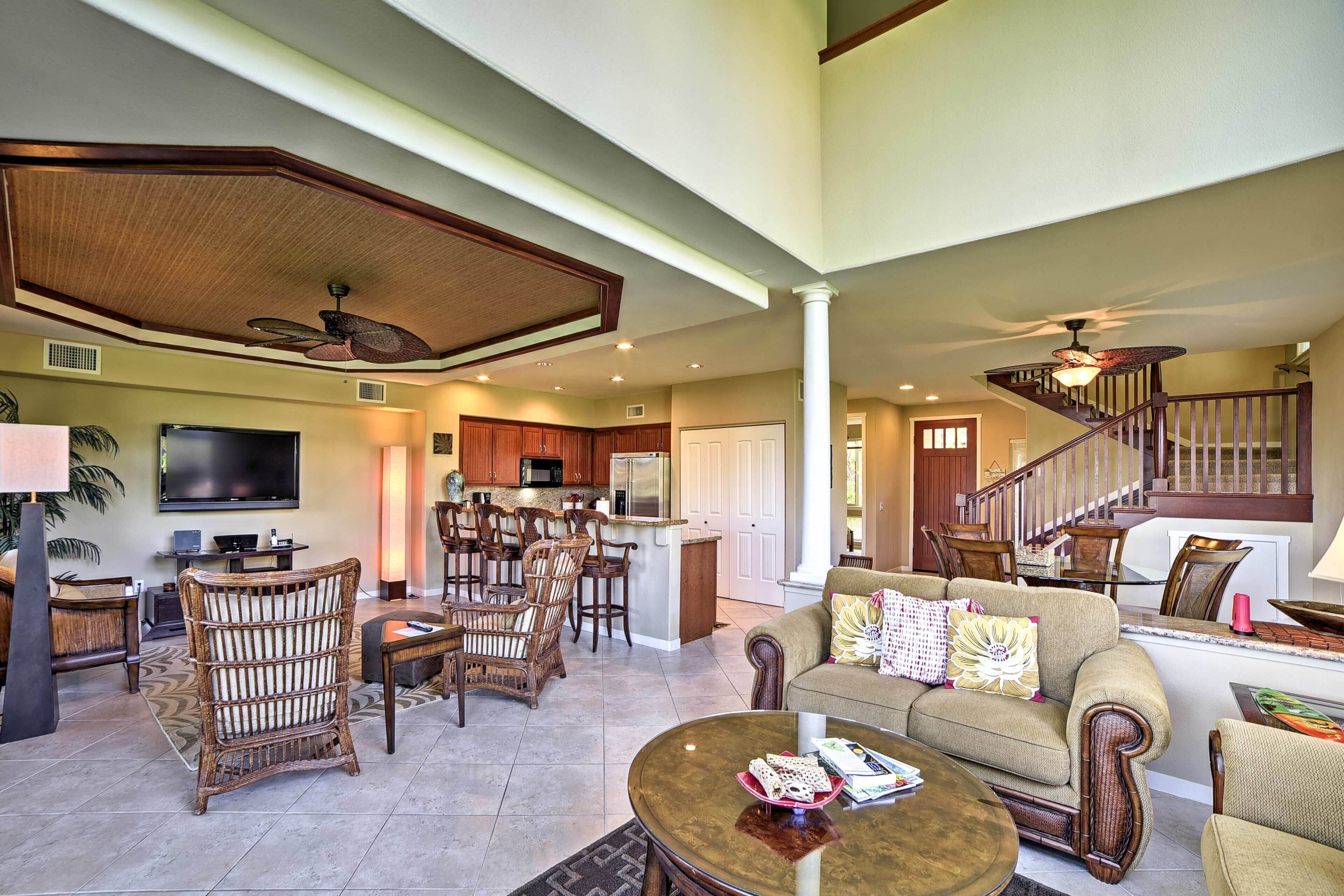 A spacious living room with a large sofa, a coffee table, and wicker chairs. There is a kitchen area with wooden cabinets and a dining table. A staircase with wooden railings leads to an upper level.