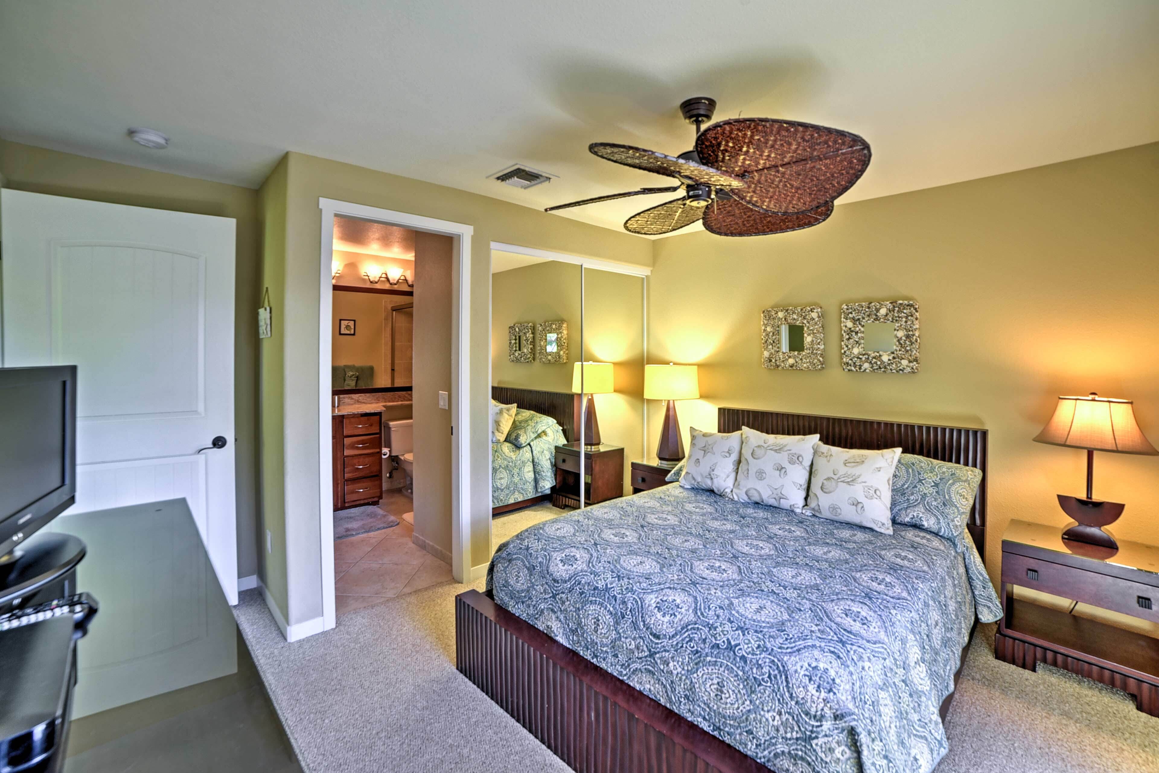 A bedroom with a bed, a ceiling fan, a TV, and a bathroom visible through an open door.