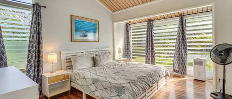 A bedroom with a bed, bedside tables, a standing fan, and large windows with blinds.