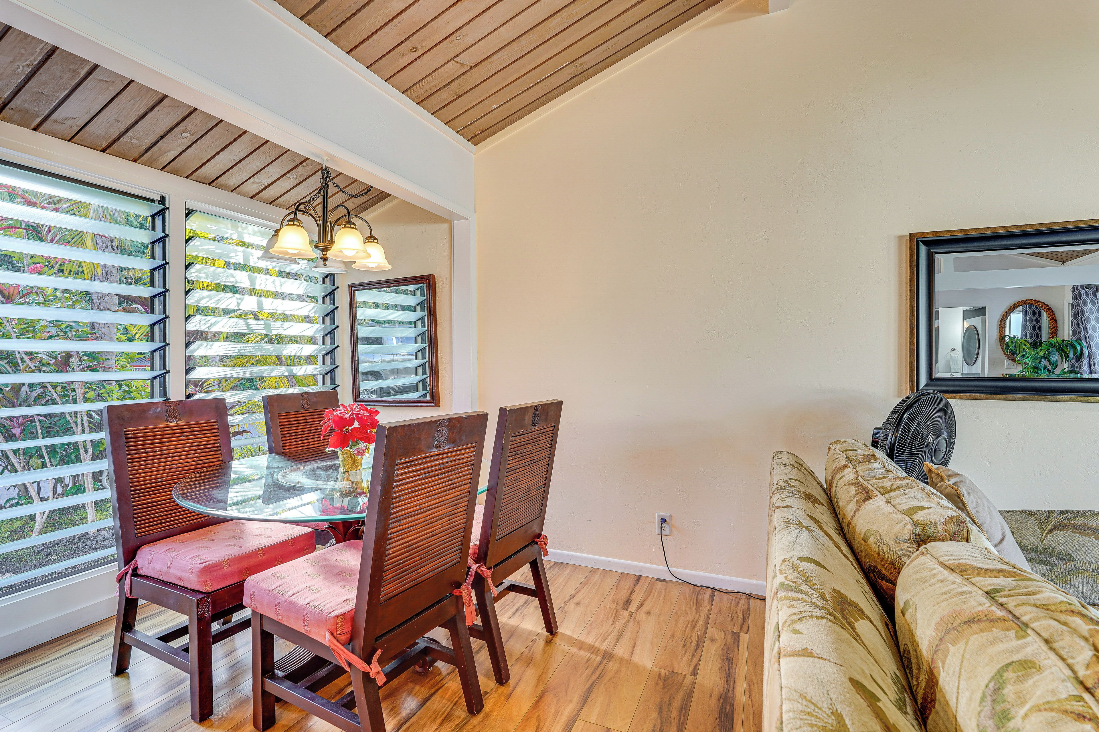A dining area with a glass table and wooden chairs, a ceiling fan, and a view through large windows.