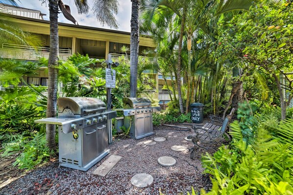 A patio area with a grill, a barbecue, and a seating bench surrounded by lush greenery and palm trees.