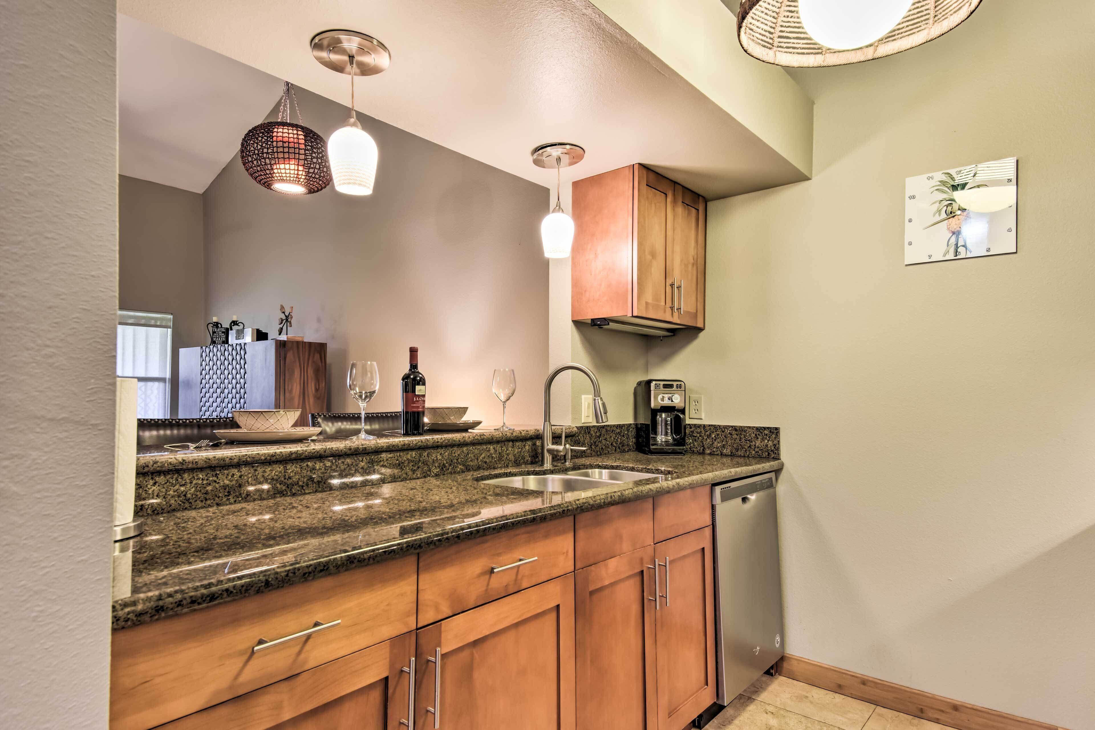 A kitchen with granite countertops, wooden cabinets, a stainless steel sink, a coffee maker, and a dishwashing unit.