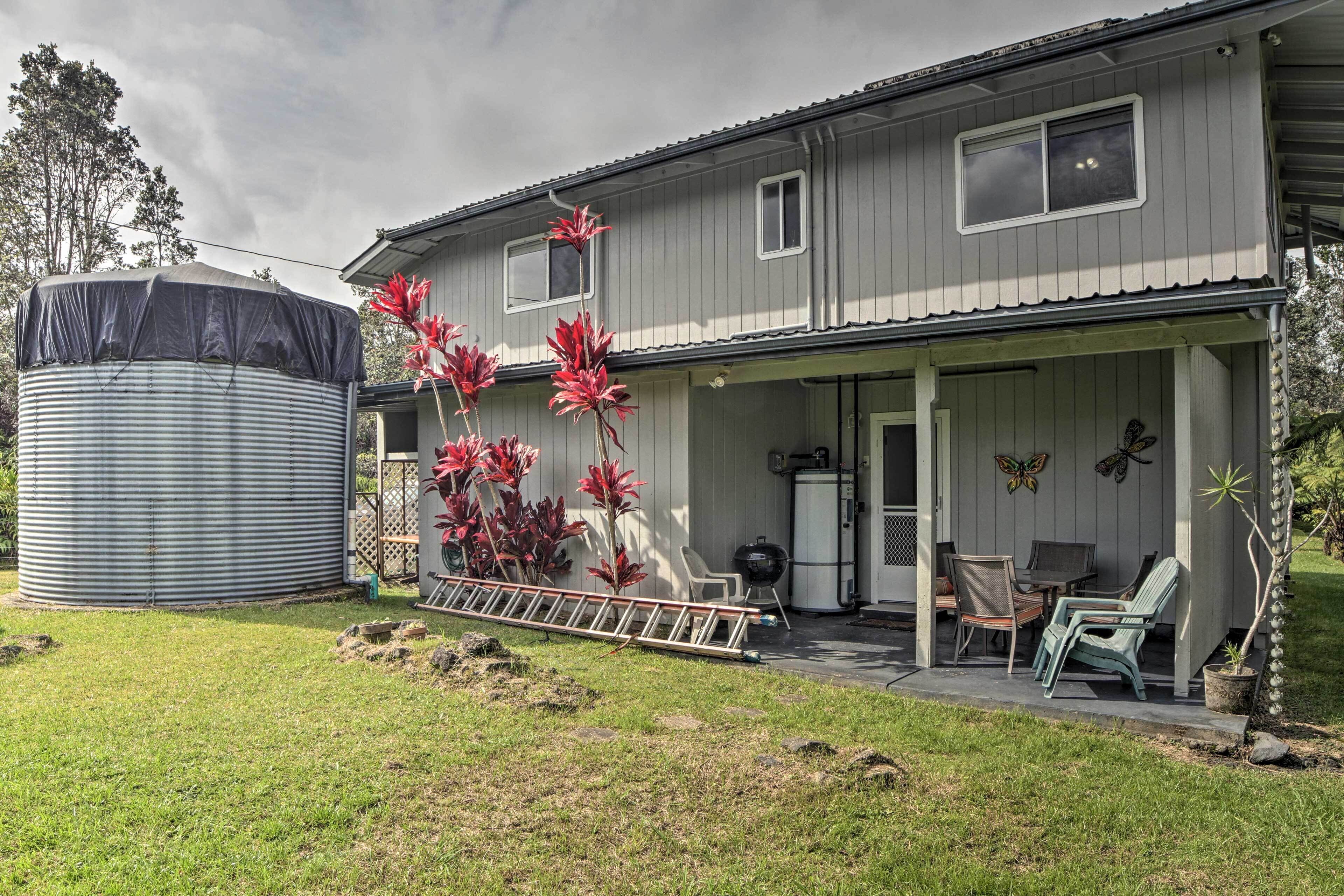 Une maison à deux étages dotée d’un porche couvert, d’un grand réservoir d’eau et de plantes à feuillage rouge.
