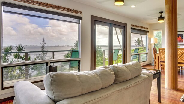 A modern living room with a large window overlooking a scenic view of the sea and palm trees. - Hilo Home w/ Private Deck + Stunning Ocean Views! (Hilo)