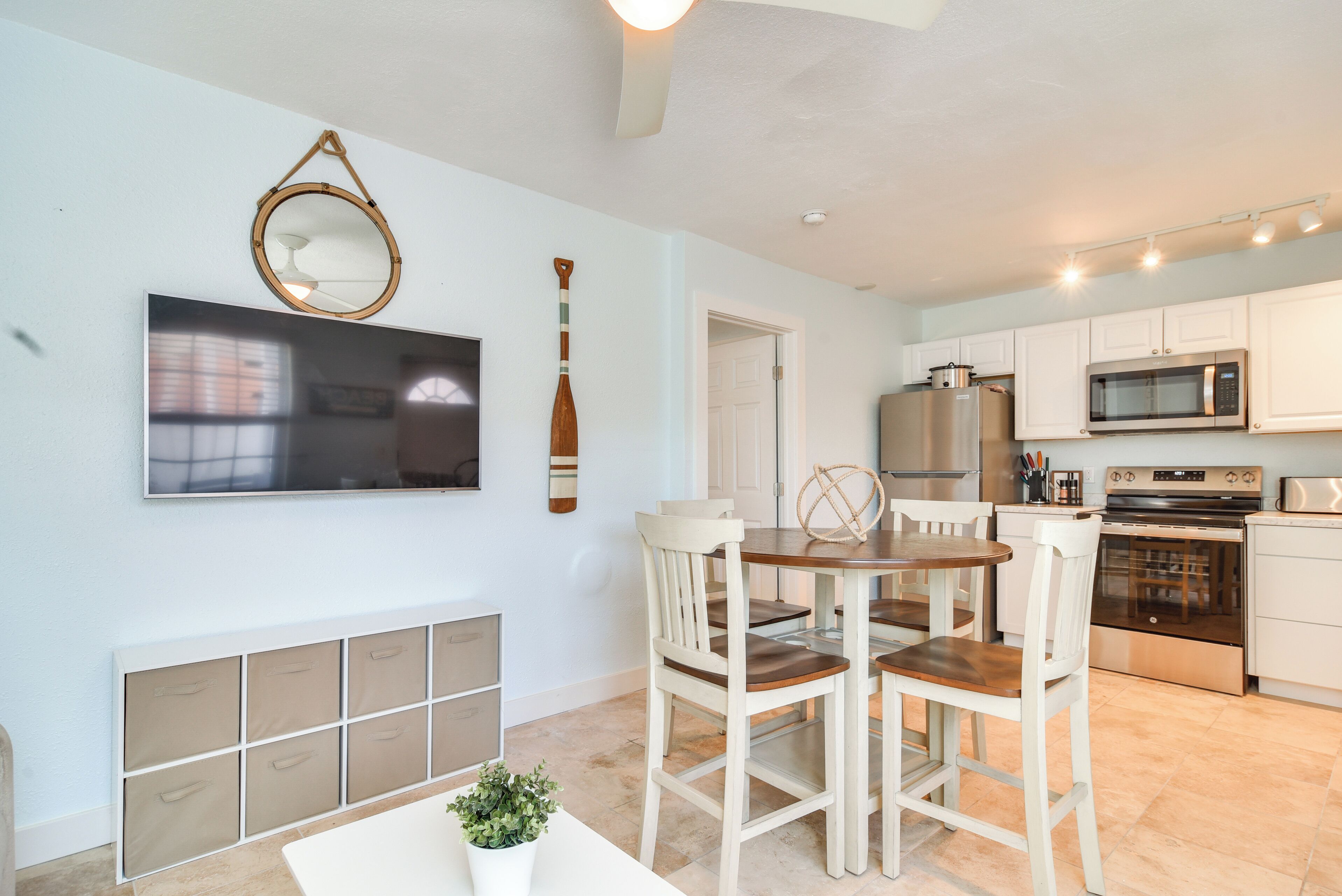 Una cocina moderna con armarios blancos, un refrigerador de acero inoxidable, un microondas y un horno. Hay un comedor con una mesa de madera y sillas blancas.