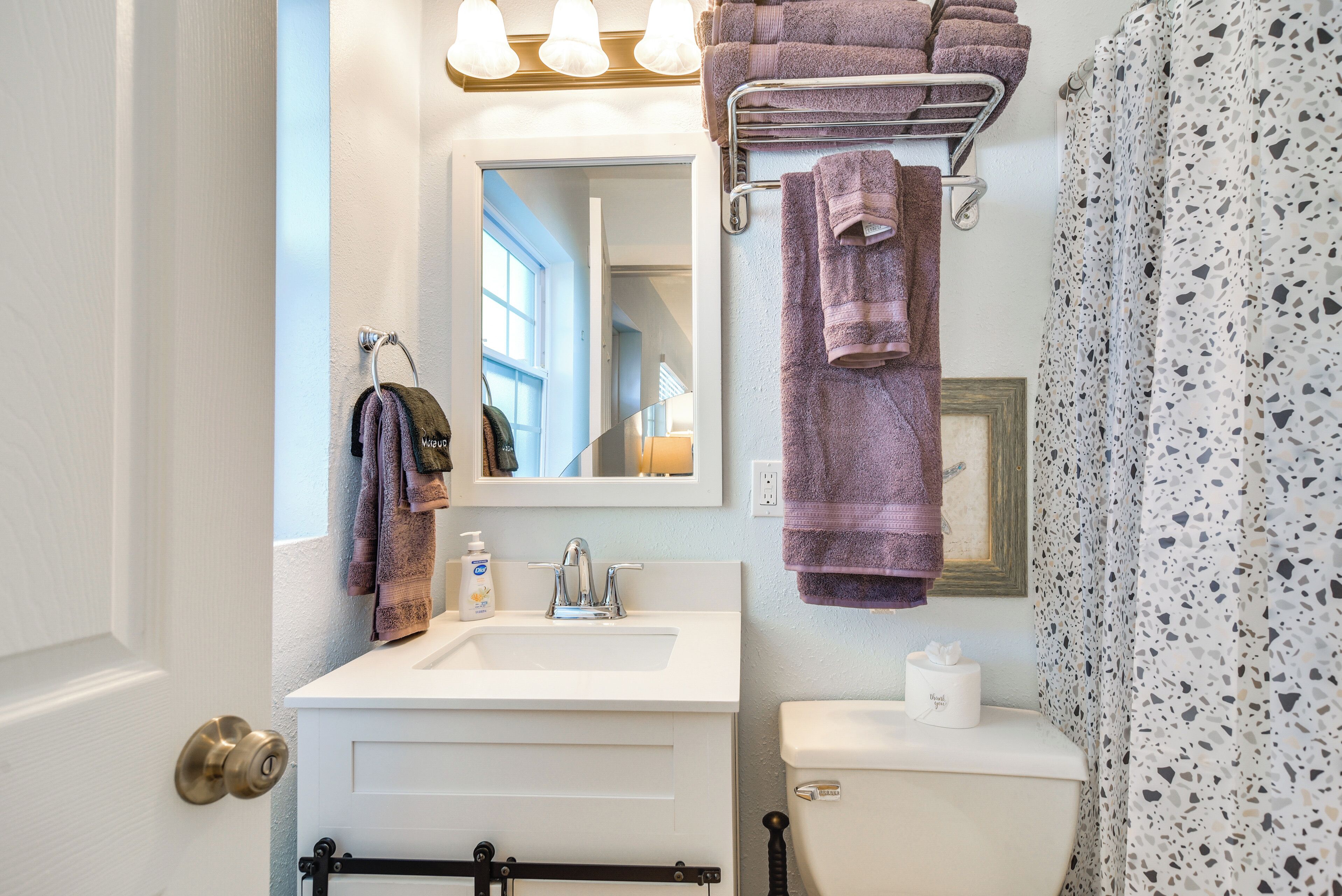 A bathroom with a white sink, a mirror, a towel rack with multiple towels, a toilet, and a shower curtain with a speckled pattern.