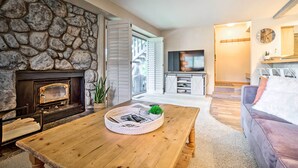 Una sala de estar con chimenea de piedra, una mesa de centro de madera con revistas, un sofá gris y un televisor al fondo.